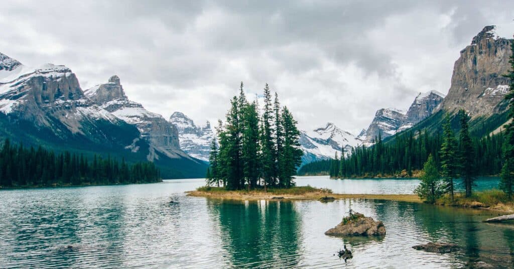 View of a river between tall snow peaked mountains and trees on a small island in the middle.