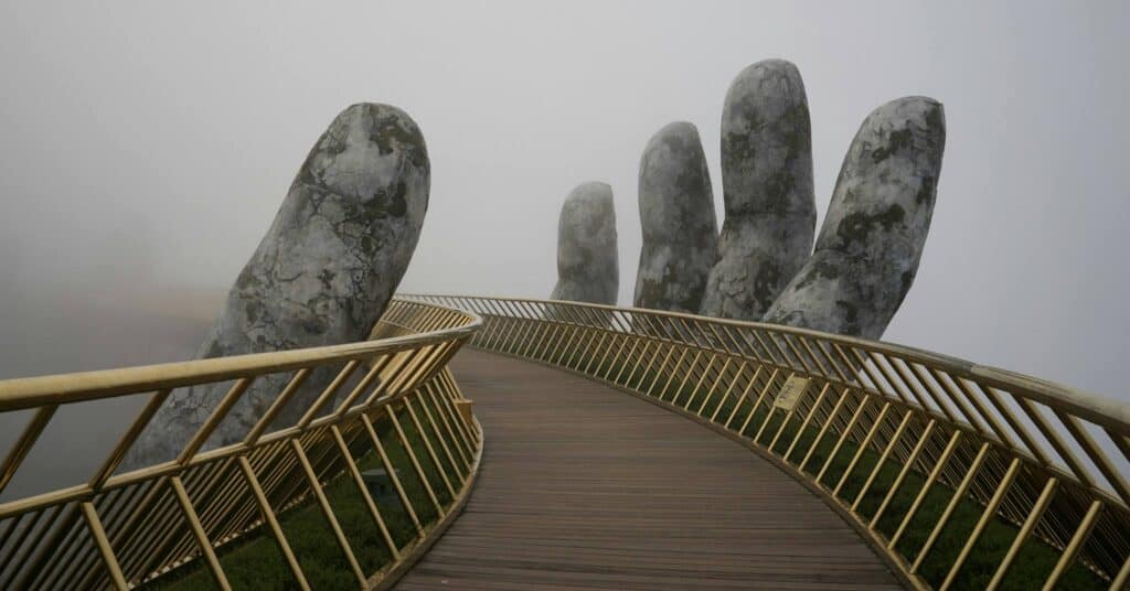 A large hand made of stone reaching out of the mist to grab onto a wooden bridge.