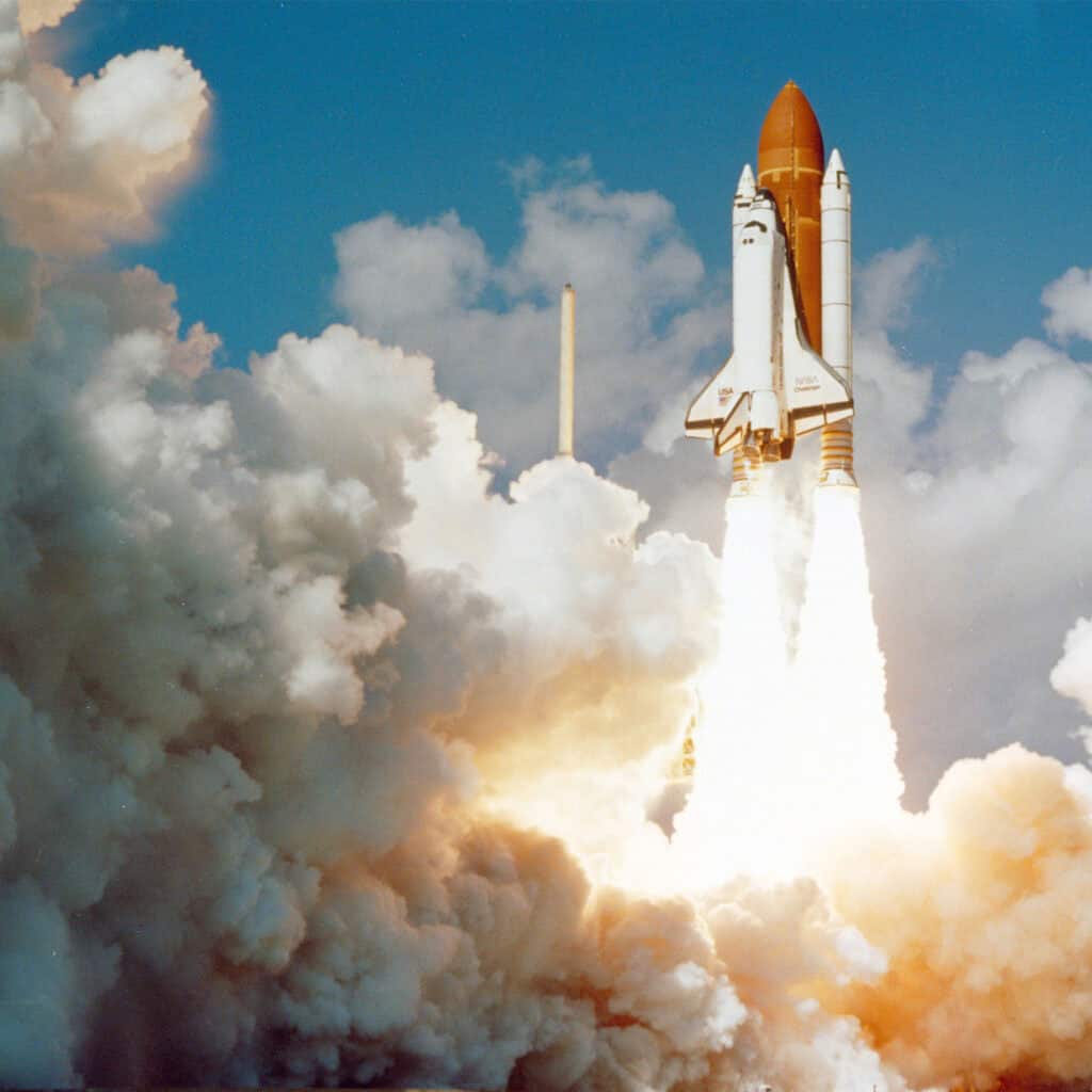 The Space Shuttle Challenger launches into a bright blue sky, leaving behind massive plumes of billowed exhaust smoke and fire.
